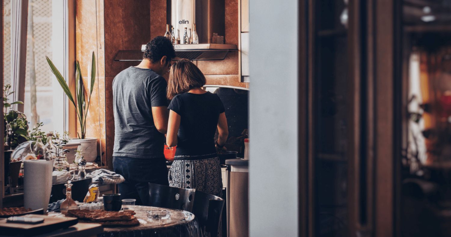 "Two People Cooking Over a Stove Facing Away From the Camera In A Kitchen