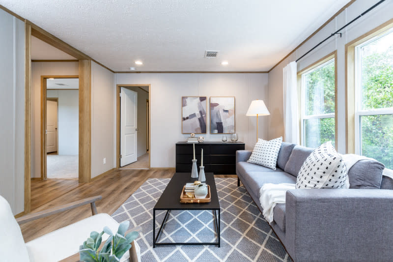 The family room in this manufactured home has ample lighting, a gray rug, and a gray couch. There are black accent pieces of furniture as well.