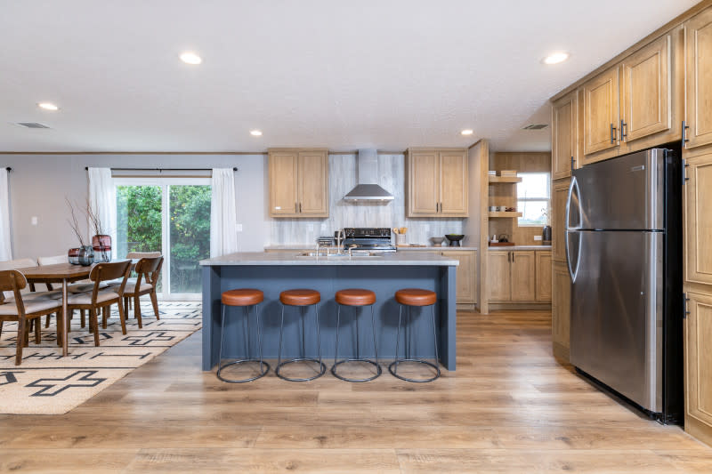 The kitchen in this Clayton Built home shows a blue kitchen island with four leather stools. Tons of light-colored wood cabinets line the walls. The refrigerator is off to the right surrounded by cabinets as well. Hardwood style floors throughout.