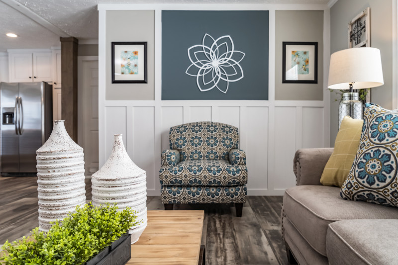 Living room of a Clayton Built® manufactured home with white wainscoting built into an accent wall with photos, a blue and yellow armchair, a beige couch and a wooden coffee table with white vases and plants.